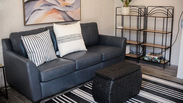 Een moderne woonkamer met een donker leren bank, gestreepte kussens, een zwart geweven poef, een zwarte metalen boekenkast en een groot abstract schilderij aan de muur.