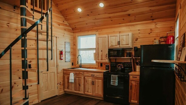 A wooden cabin interior with a kitchen area, wooden cabinets, a black stove, and a black refrigerator.