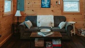 A cozy cabin interior with a wooden ceiling, a ceiling fan, a fireplace, a sofa, a coffee table, and a window with blinds.