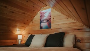 A cozy wooden cabin bedroom with a bed, pillows, a nightstand with a lamp, and a framed picture of a cherry blossom bridge.