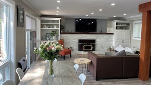 A modern living room with a fireplace, a television, and a dining area with a vase of flowers. - Elegant waterfront retreat on Paudash Lake with sunset views! (Highlands east)