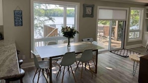 A dining area with a large table and chairs, a vase of flowers, and a view of a lake through large windows. - Elegant waterfront retreat on Paudash Lake with sunset views! (Highlands east)