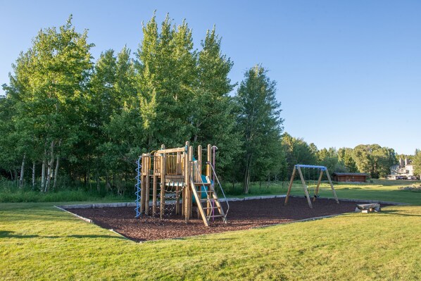 Un parque infantil con una estructura de madera para escalar y columpios, rodeado de césped y árboles.