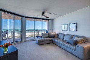 En moderne stue med en stor grå hjørnesofa, en glasdør der fører ud til en balkon med havudsigt, og en loftsvifte.