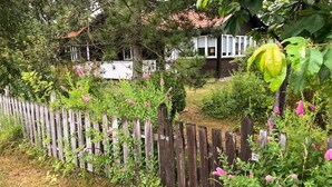 Una recinzione in legno con un giardino in primo piano, una casa con tetto rosso sullo sfondo e un edificio bianco con tetto scuro.
