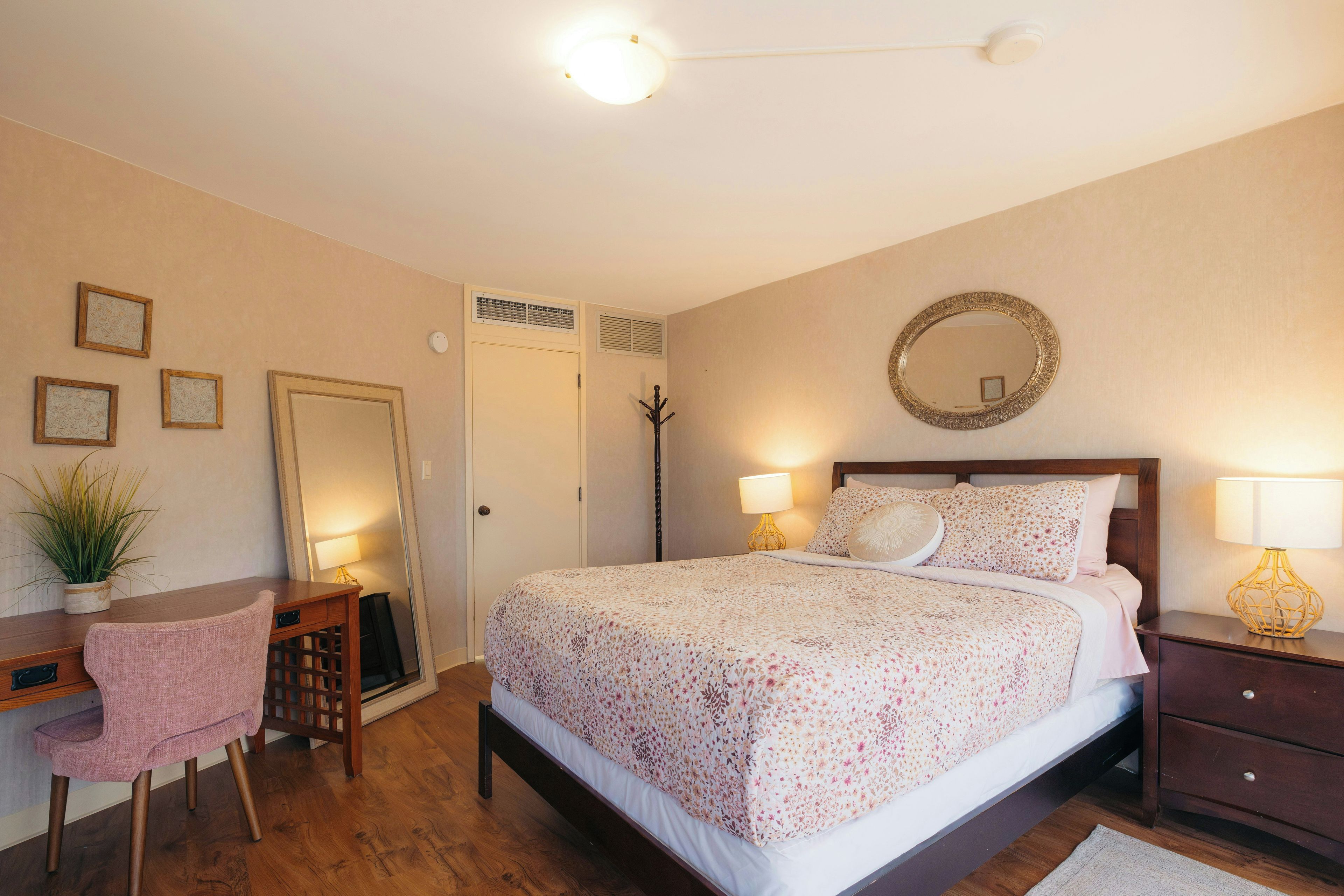 A bedroom with a bed, bedside lamps, a desk, a chair, a mirror, and a potted plant.