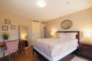 A bedroom with a bed, bedside lamps, a desk, a chair, a mirror, and a potted plant.
