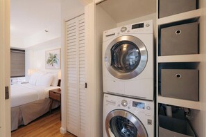 A laundry room with two stacked front-loading washing machines and a bed with a nightstand in the adjacent bedroom. - Bishop Suites Downtown Honolulu (Honolulu)