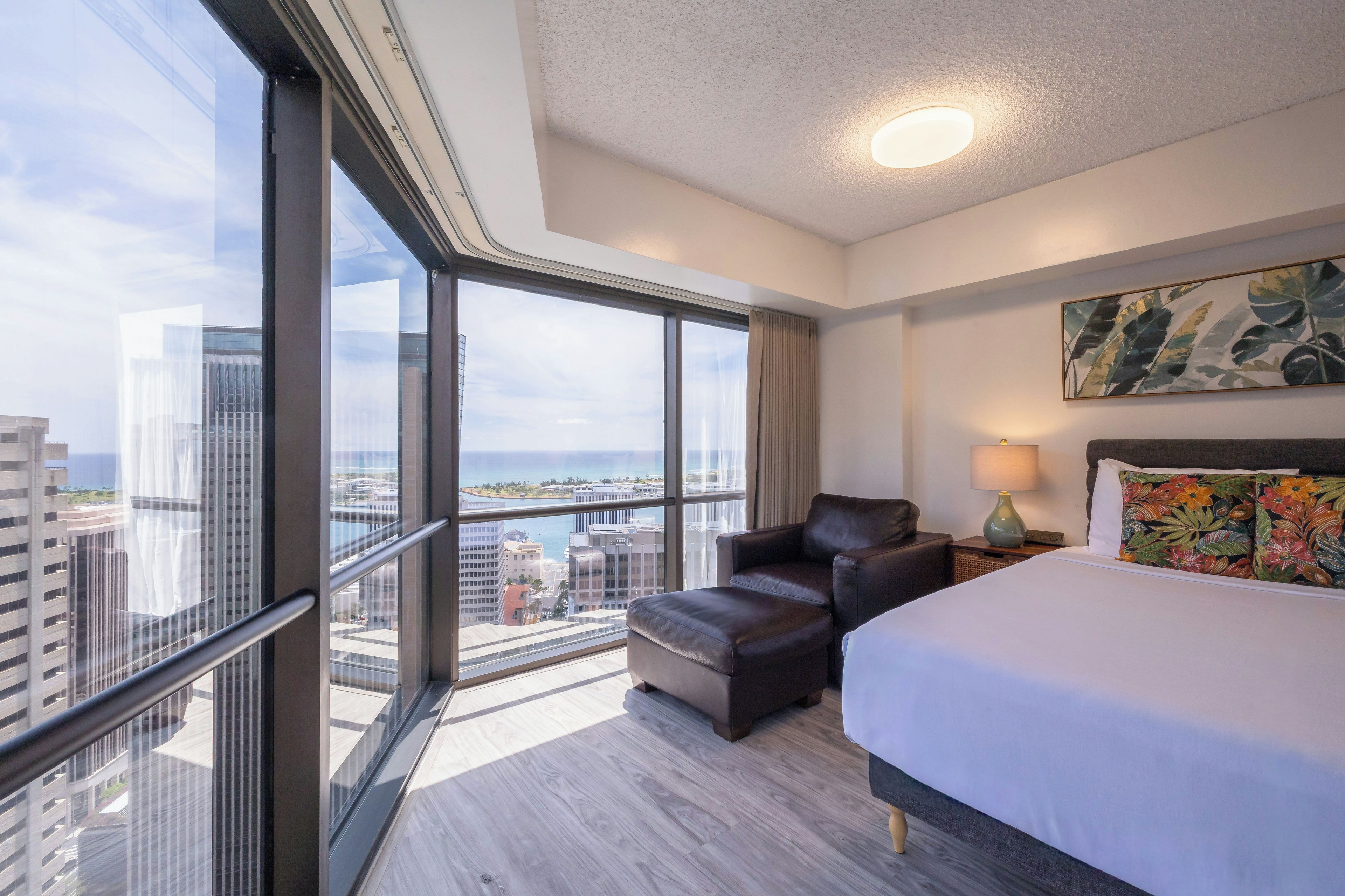 A hotel room with a large window, a bed, a chair, a nightstand, and a painting on the wall.