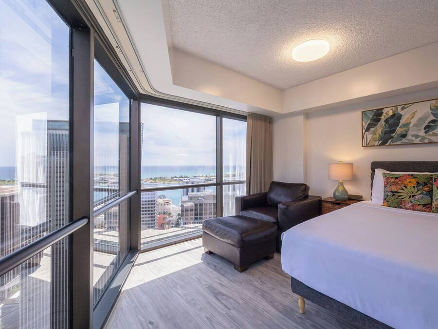 A hotel room with a large window, a bed, a chair, a nightstand, and a painting on the wall.