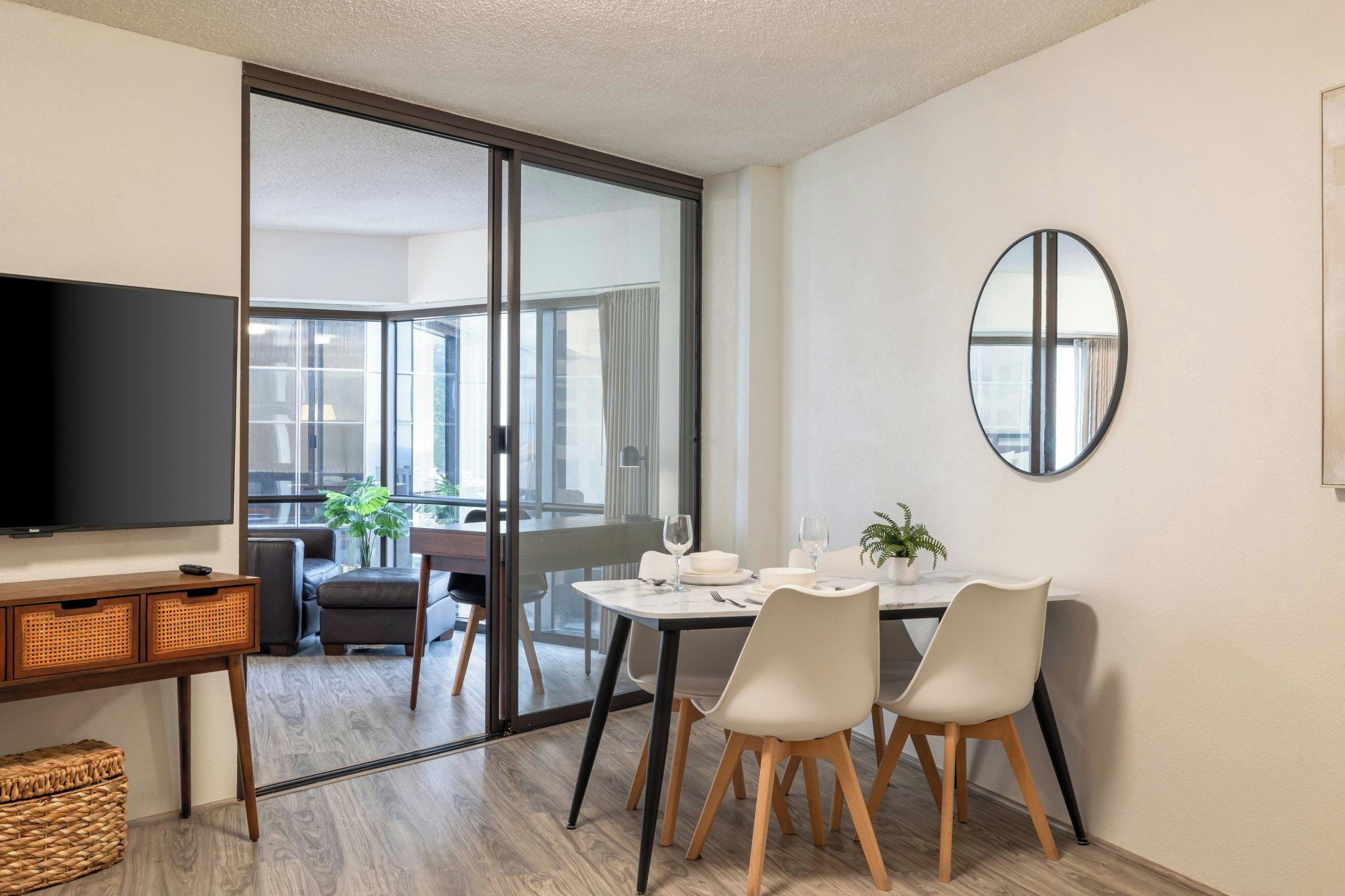 Un soggiorno moderno con una TV a schermo piatto, un tavolo da pranzo con sedie, un divano e una porta vetrata scorrevole che dà su un balcone.