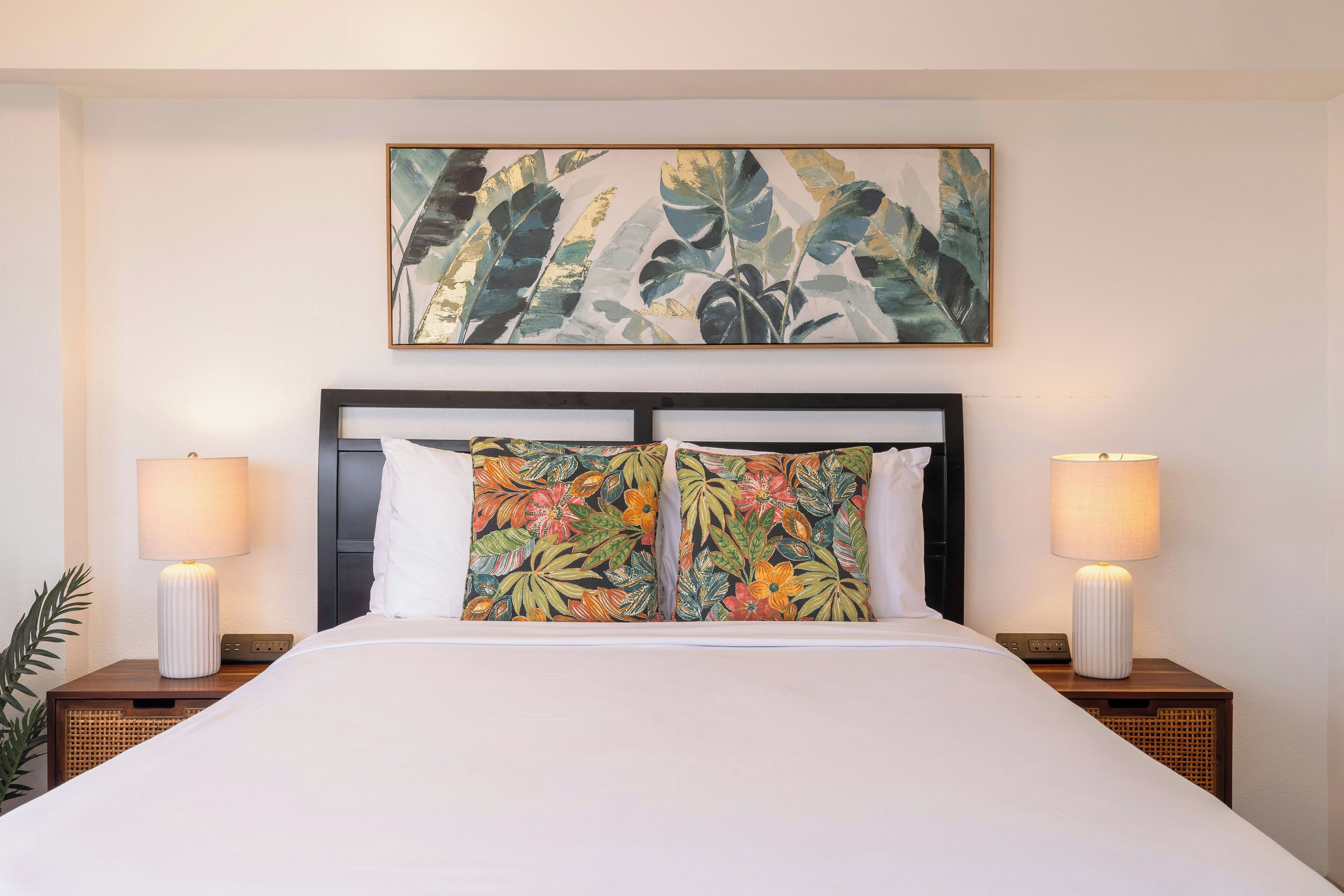 A hotel room with a large bed, two bedside lamps, a headboard with a floral pillow, and a tropical-themed painting above the bed.