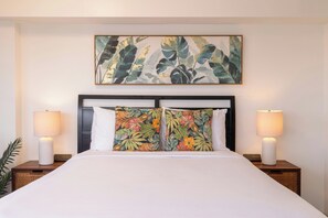 Une chambre d’hôtel avec un grand lit, deux lampes de chevet, une tête de lit agrémentée d’un coussin à motifs floraux et un tableau sur le thème des pays tropicaux au-dessus du lit.