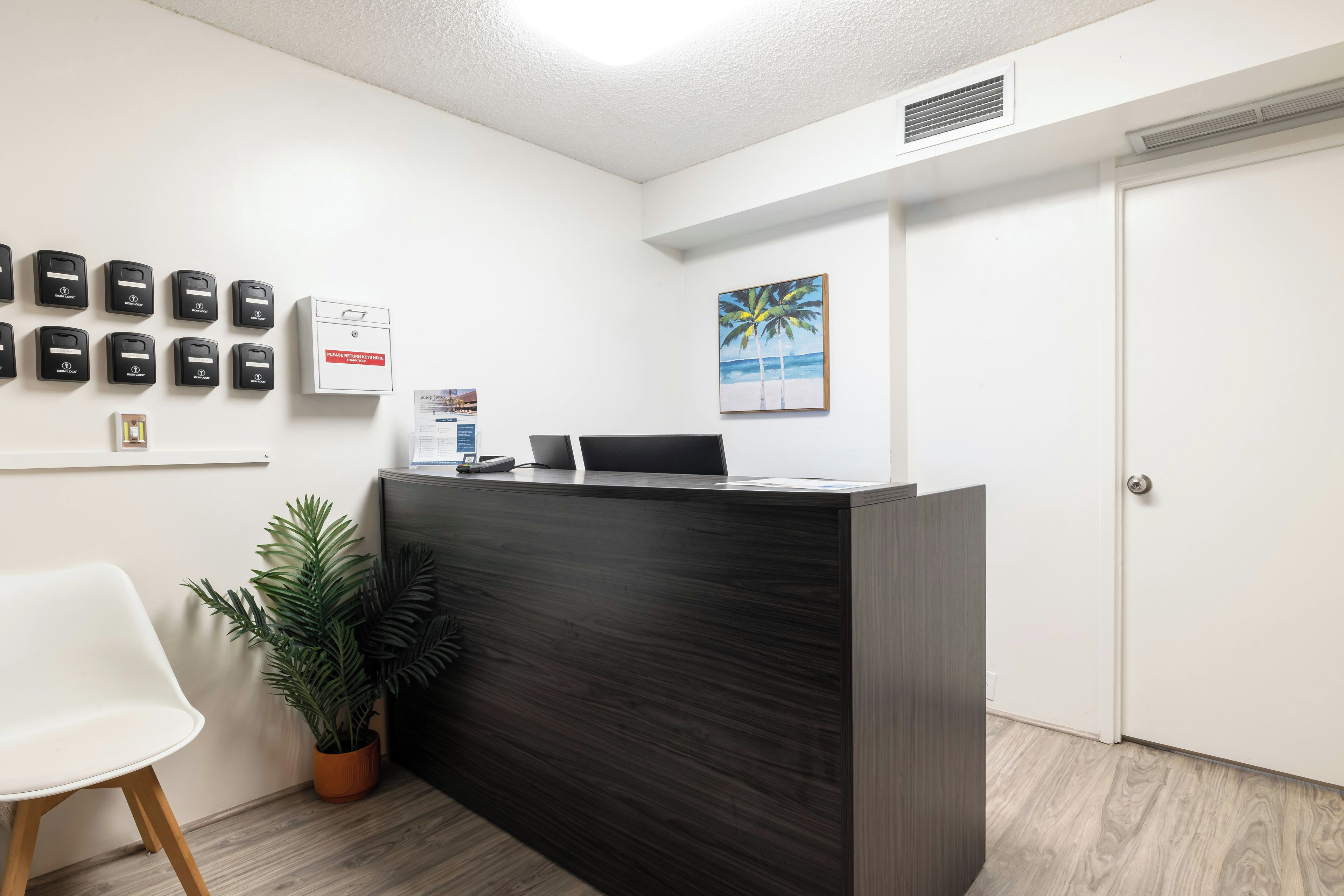 A reception desk with a computer, a plant, and a painting of palm trees.