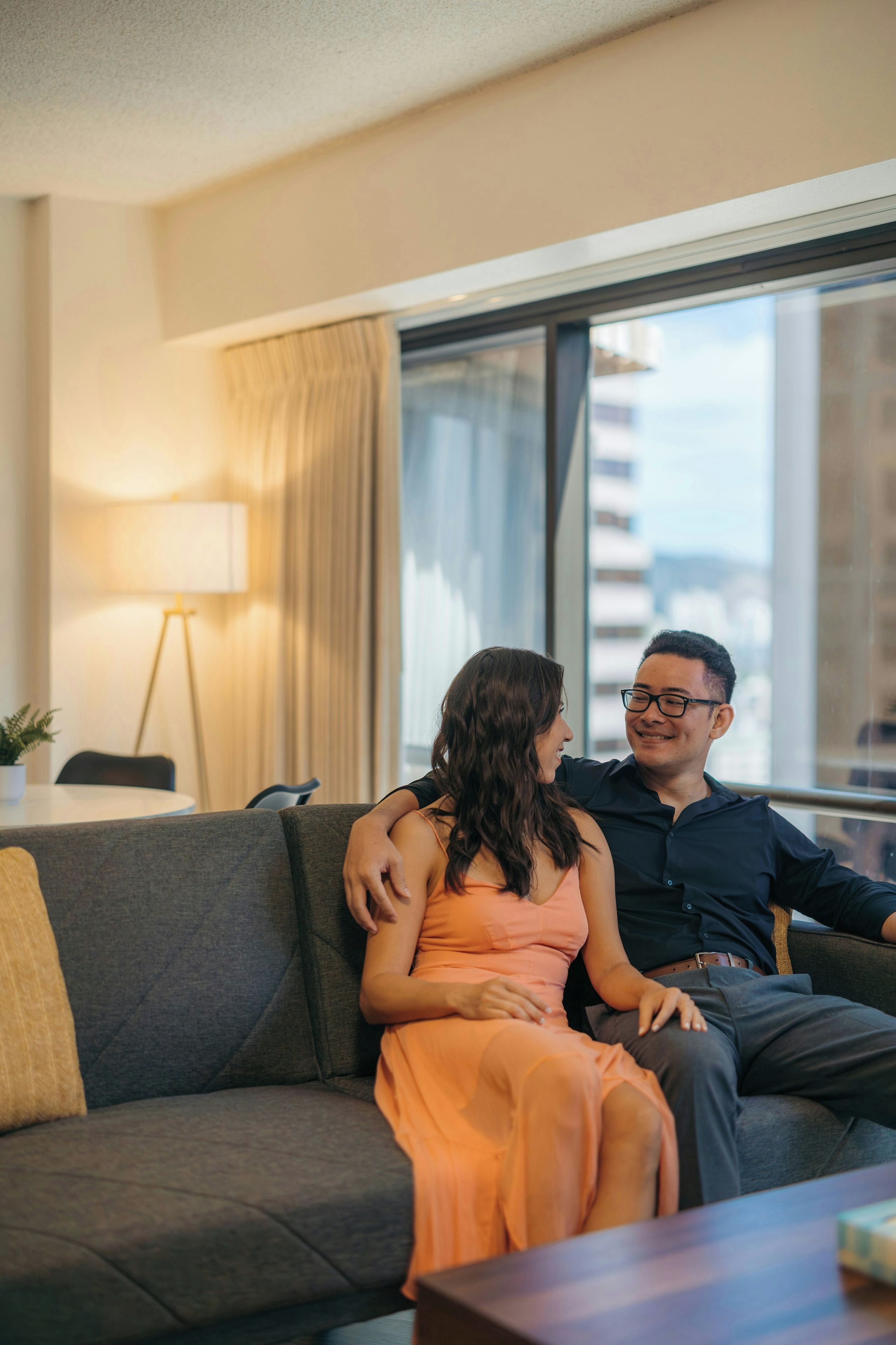 Una pareja sentada en un sofá gris en una sala de estar moderna con un ventanal grande, una lámpara y vistas a edificios.