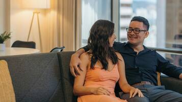 Una pareja sentada en un sofá gris en una sala de estar moderna con un ventanal grande, una lámpara y vistas a edificios.