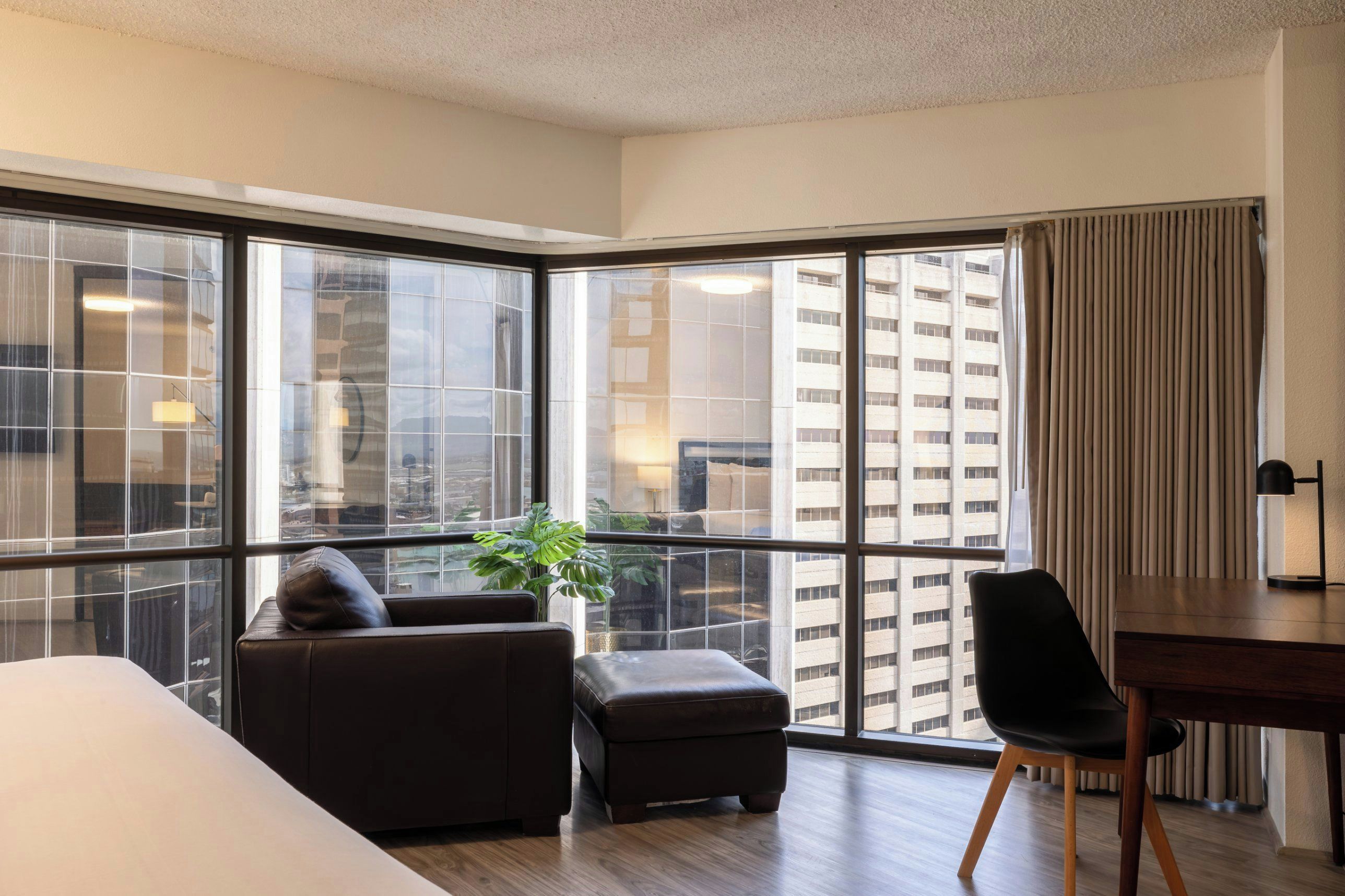 A modern hotel room with a large corner window, a leather sofa, a small ottoman, a dining table with a chair, and a potted plant.