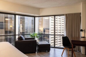 A modern hotel room with a large corner window, a leather sofa, a small ottoman, a dining table with a chair, and a potted plant. - Bishop Suites Downtown Honolulu (Honolulu)
