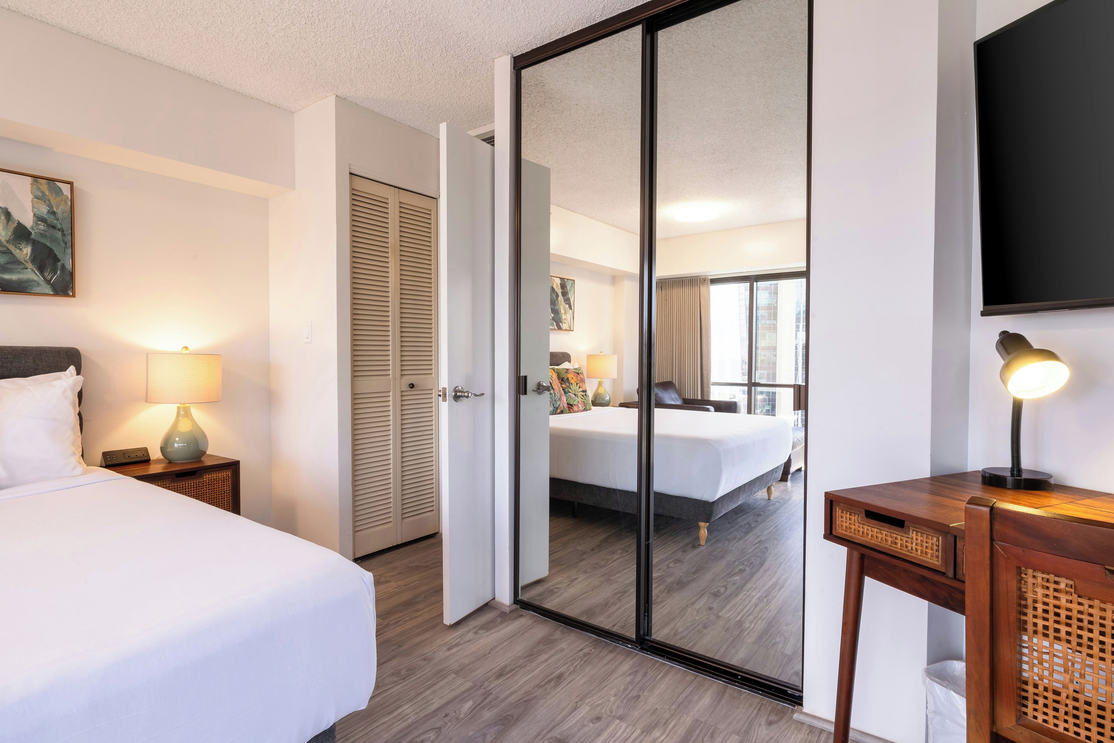 Una habitación de hotel con una cama, mesitas de noche, un escritorio, un espejo y un televisor.