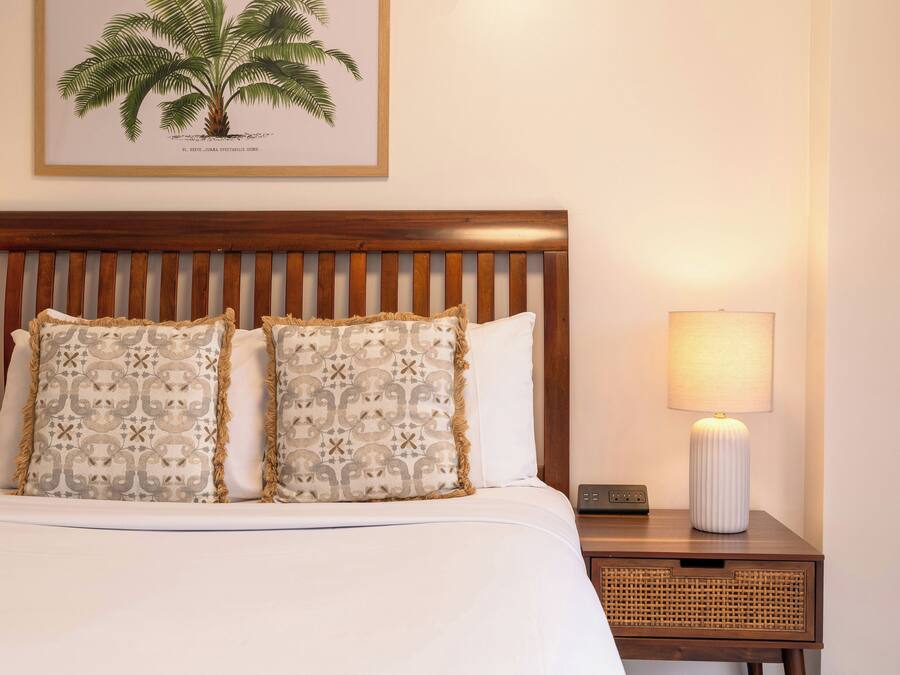A neatly made bed with patterned pillows, a wooden headboard, a bedside lamp, and a framed botanical illustration on the wall.