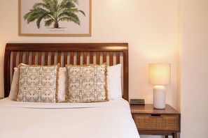 A neatly made bed with patterned pillows, a wooden headboard, a bedside lamp, and a framed botanical illustration on the wall. - Bishop Suites Downtown Honolulu (Honolulu)