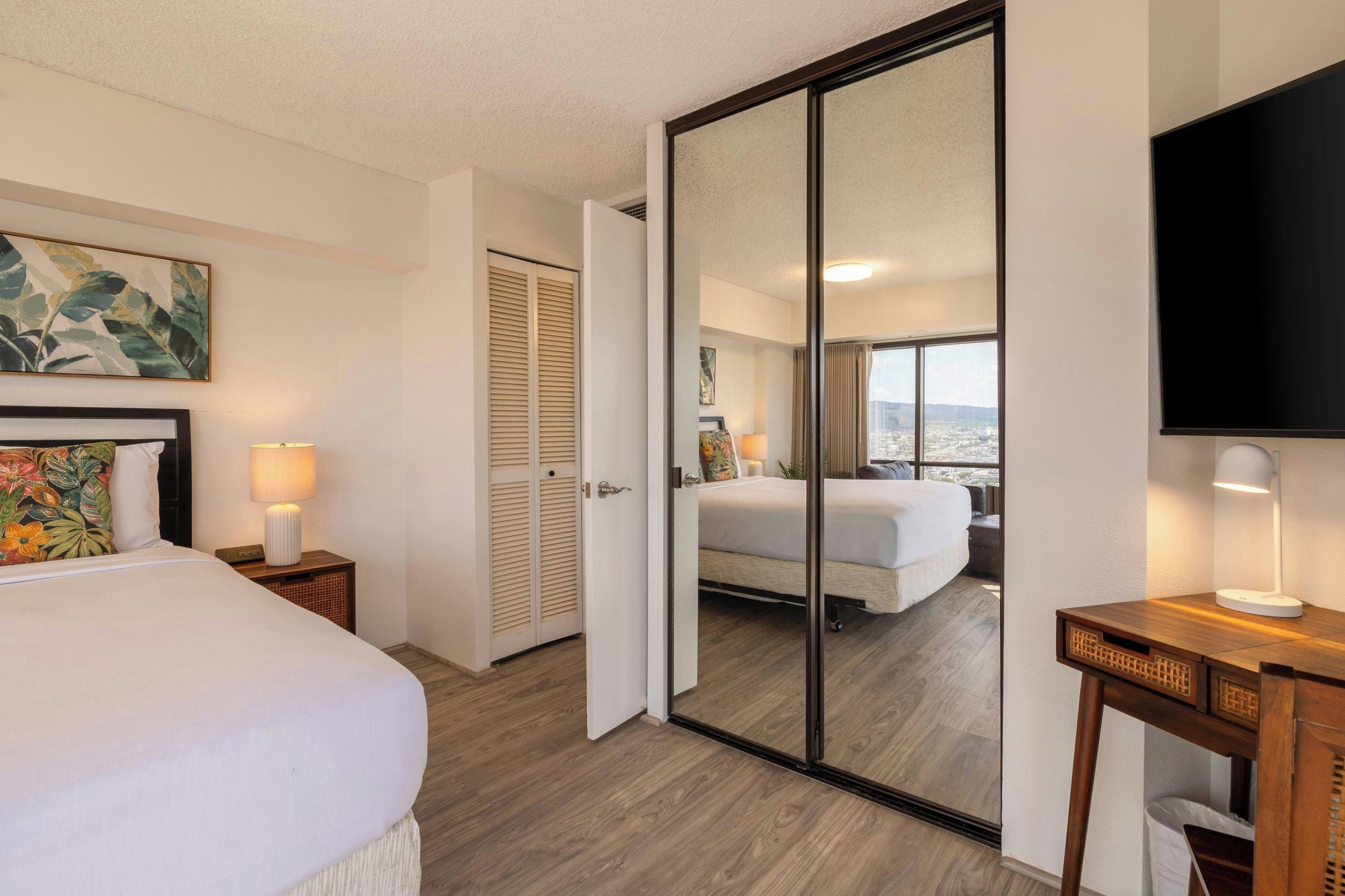 Habitación de hotel con cama, escritorio, silla, televisión, ventana con vista a la ciudad y puerta corrediza.