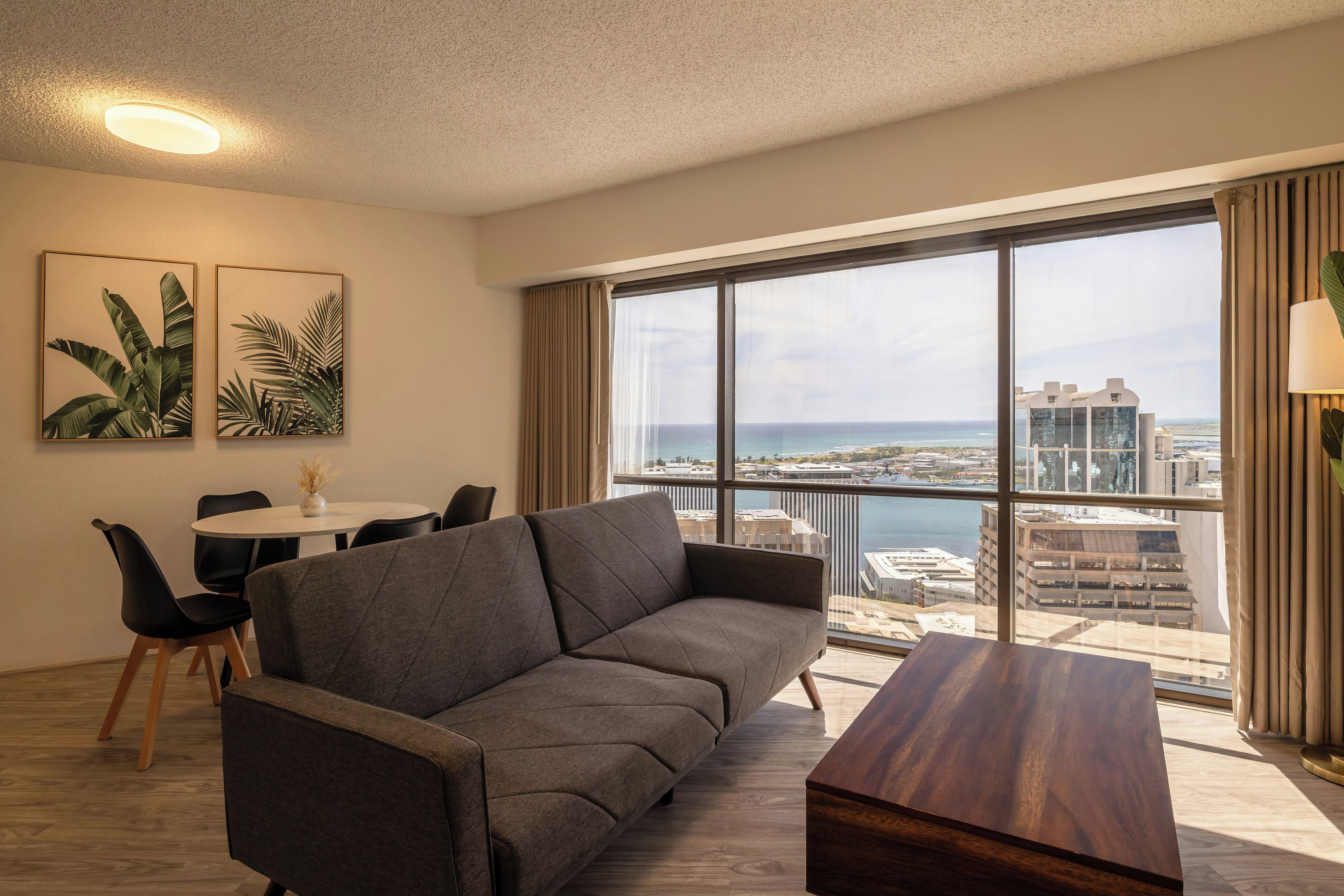 A modern living room with a grey sofa, a wooden coffee table, and a dining area with a white table and black chairs. Large windows offer a view of the city and the sea.
