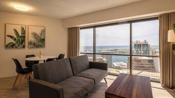 A modern living room with a grey sofa, a wooden coffee table, and a dining area with a white table and black chairs. Large windows offer a view of the city and the sea.