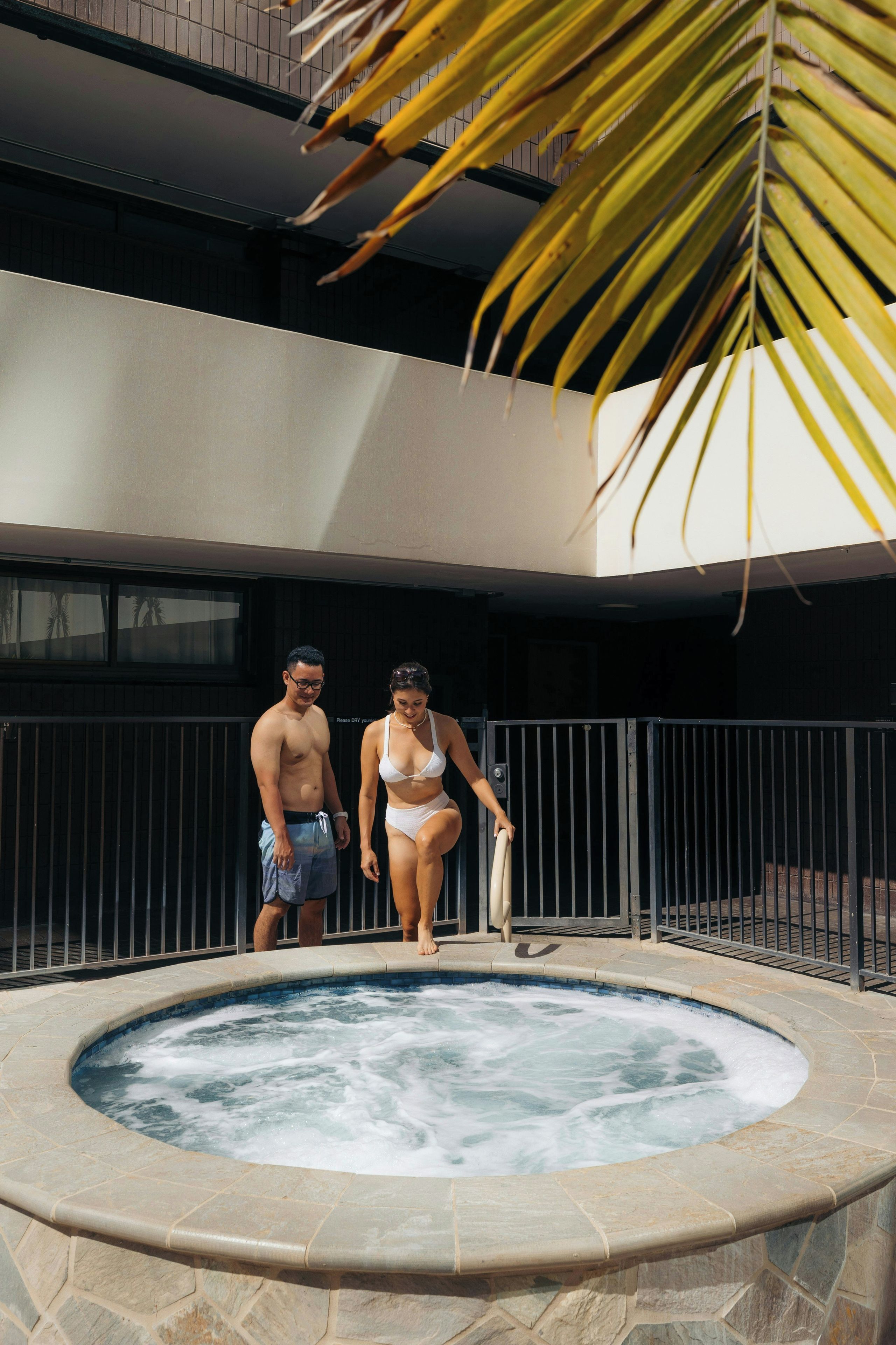 A hot tub with a stone border, a metal fence, and a building with a balcony in the background.
