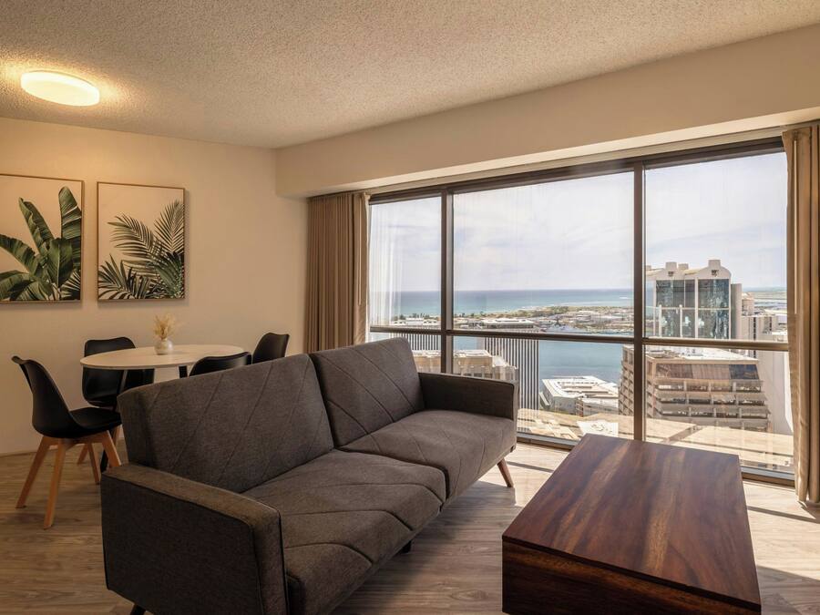 Ein modernes Wohnzimmer mit einem grauen Sofa, einem Holztisch und einem Essbereich mit weißem Tisch und schwarzen Stühlen. An der Wand befinden sich zwei gerahmte Pflanzenbilder, und ein großes Fenster bietet Blick auf die Stadt und das Meer.
