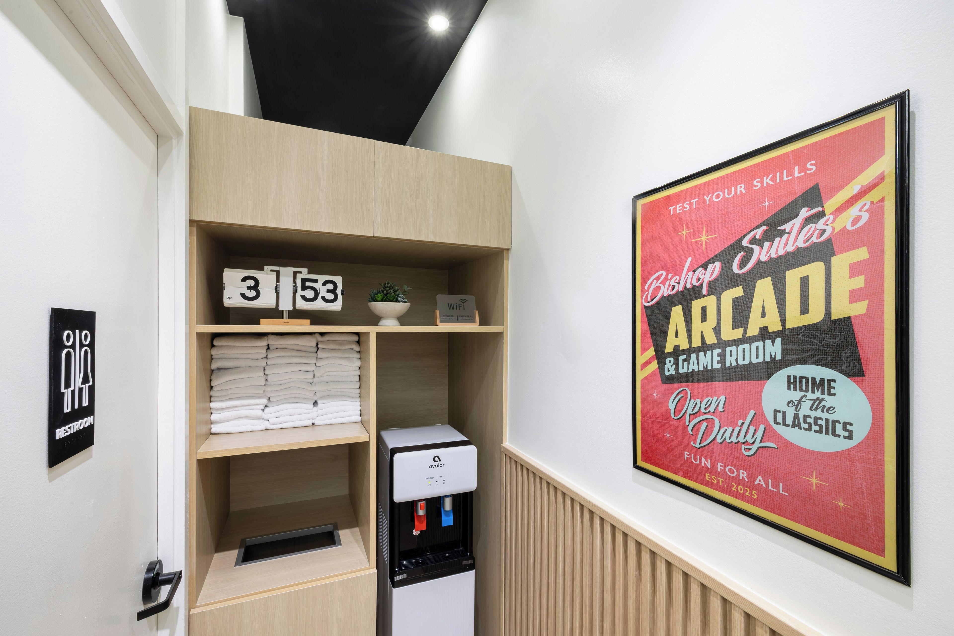 Habitación de hotel con estante empotrado que contiene toallas dobladas, un reloj y un cartel de Wi-Fi. Se observa un cartel enmarcado que anuncia una sala de juegos.