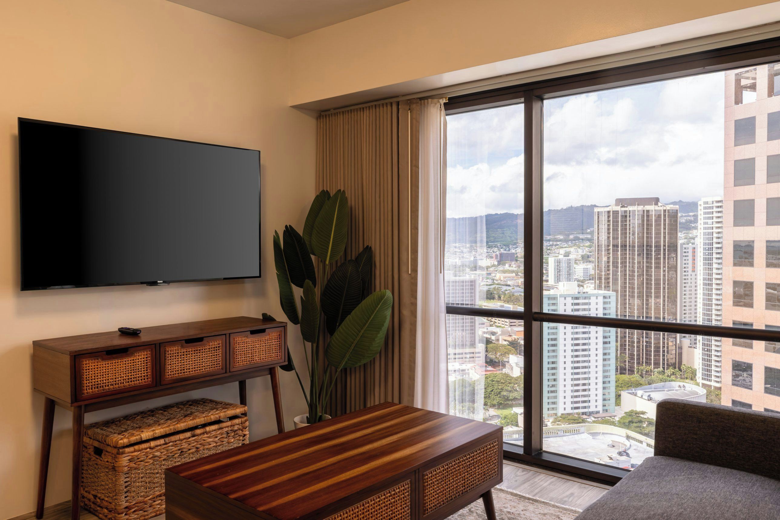 Habitación de hotel moderna con un amplio ventanal que ofrece vistas a la ciudad, un televisor de pantalla plana montado en la pared, una mesita de madera con una cesta tejida y una planta grande.