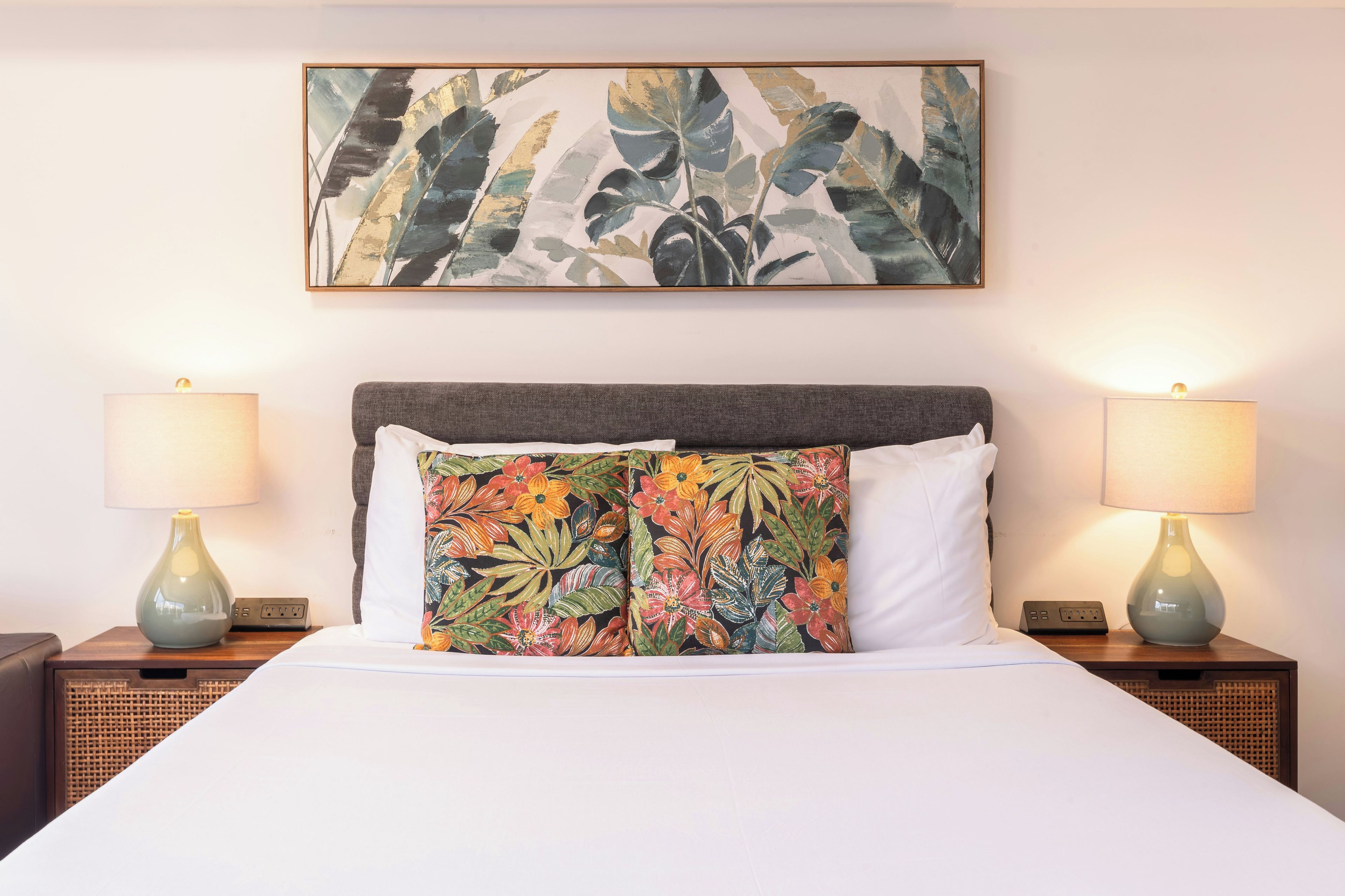 A hotel room with a bed, two nightstands, two lamps, a floral artwork above the bed, and a power strip on each nightstand.