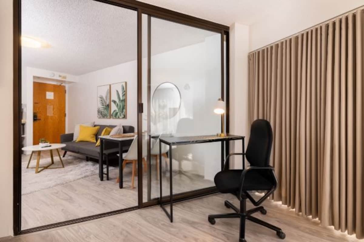 Una habitación moderna con separador de vidrio, un escritorio, una silla de oficina negra, un sofá con cojines amarillos y una puerta de madera.