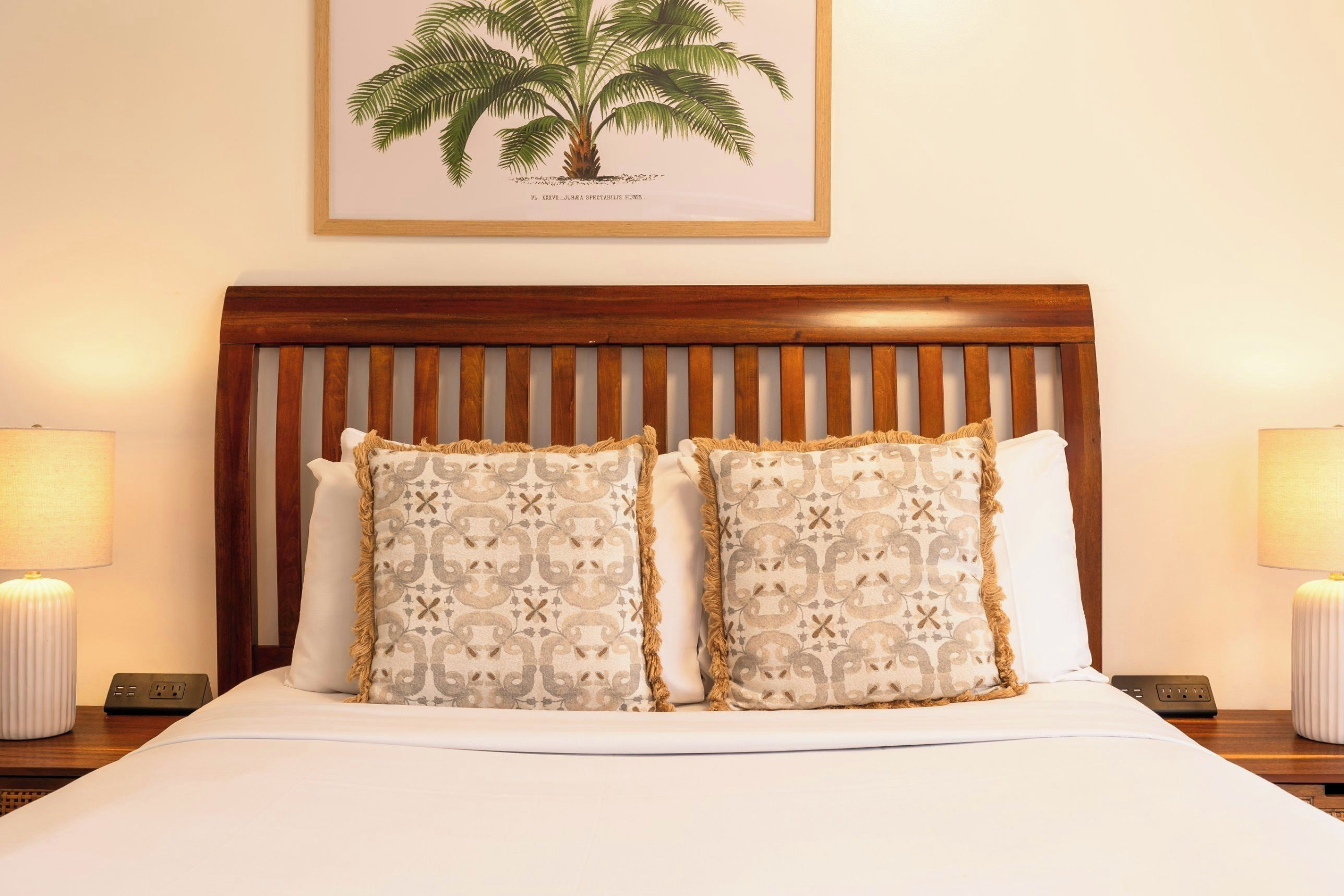 A neatly made bed with two patterned pillows, a wooden headboard, and a framed botanical illustration above.