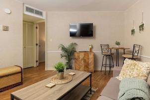 A modern living room with a wooden coffee table, a sofa, a TV mounted on the wall, and a dining area with a small table and chairs.