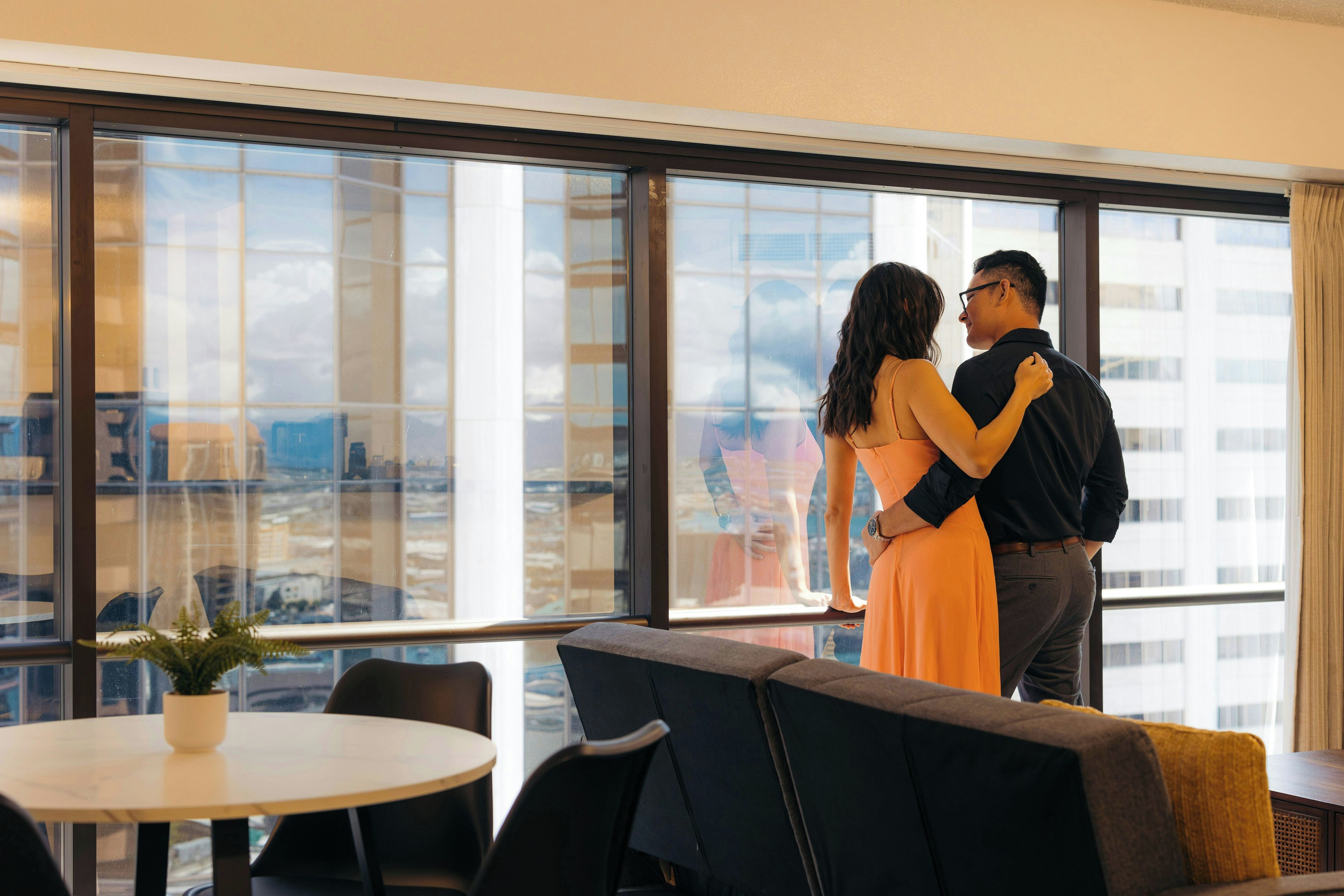 Una pareja de pie junto a un amplio ventanal con vistas a un paisaje urbano.