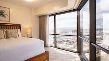 Habitación de hotel con un ventanal grande, una cama con almohadas estampadas, una lámpara de noche y vistas a un paisaje urbano.