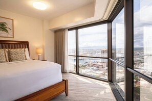 Une chambre d’hôtel avec une grande fenêtre, un lit agrémenté d’oreillers à motifs, une lampe de chevet et une vue sur le paysage urbain.