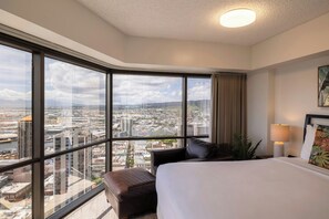 A hotel room with a large window offering a city view, a bed, a bedside table with a lamp, and a brown leather chair. - Bishop Suites Downtown Honolulu (Honolulu)