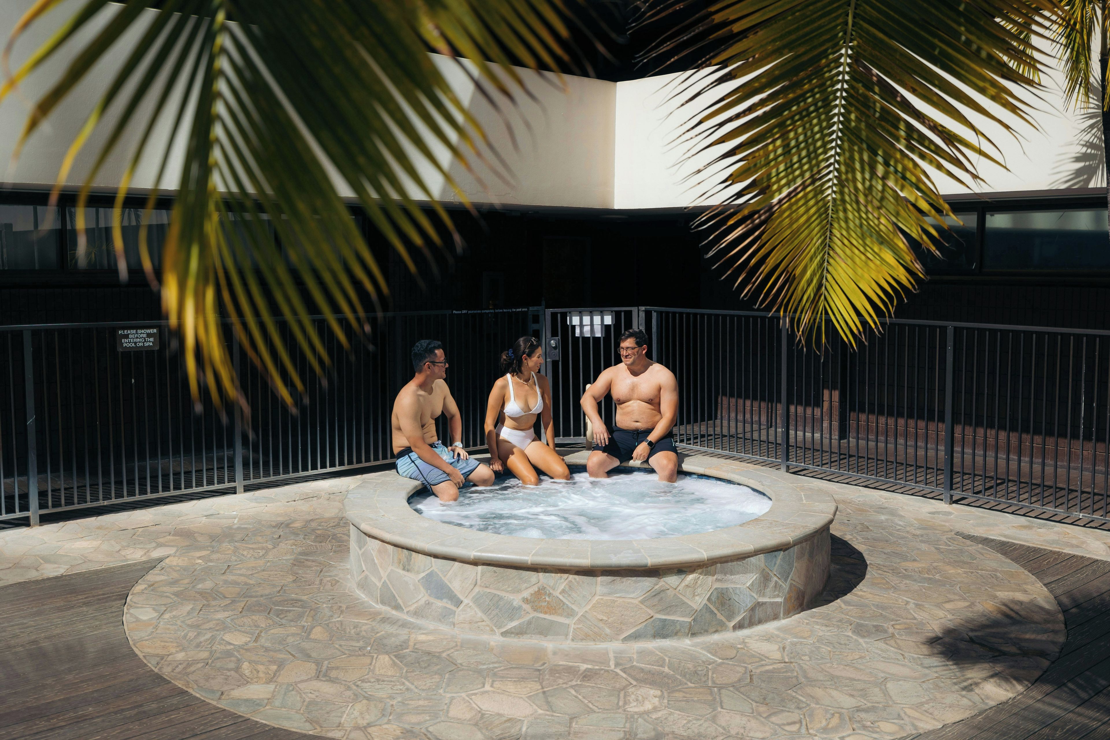 A hot tub with three people sitting around it, surrounded by stone tiles and a metal fence.