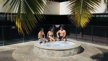 A hot tub with three people sitting around it, surrounded by stone tiles and a metal fence.