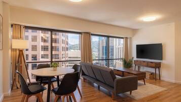 Een moderne woonkamer met een bank, eettafel, stoelen, een flatscreen televisie en grote ramen met uitzicht op de stad.