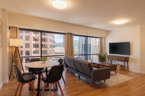 A modern living room with a sofa, dining table, chairs, a flat-screen TV, and large windows offering a city view. - Bishop Suites Downtown Honolulu (Honolulu)