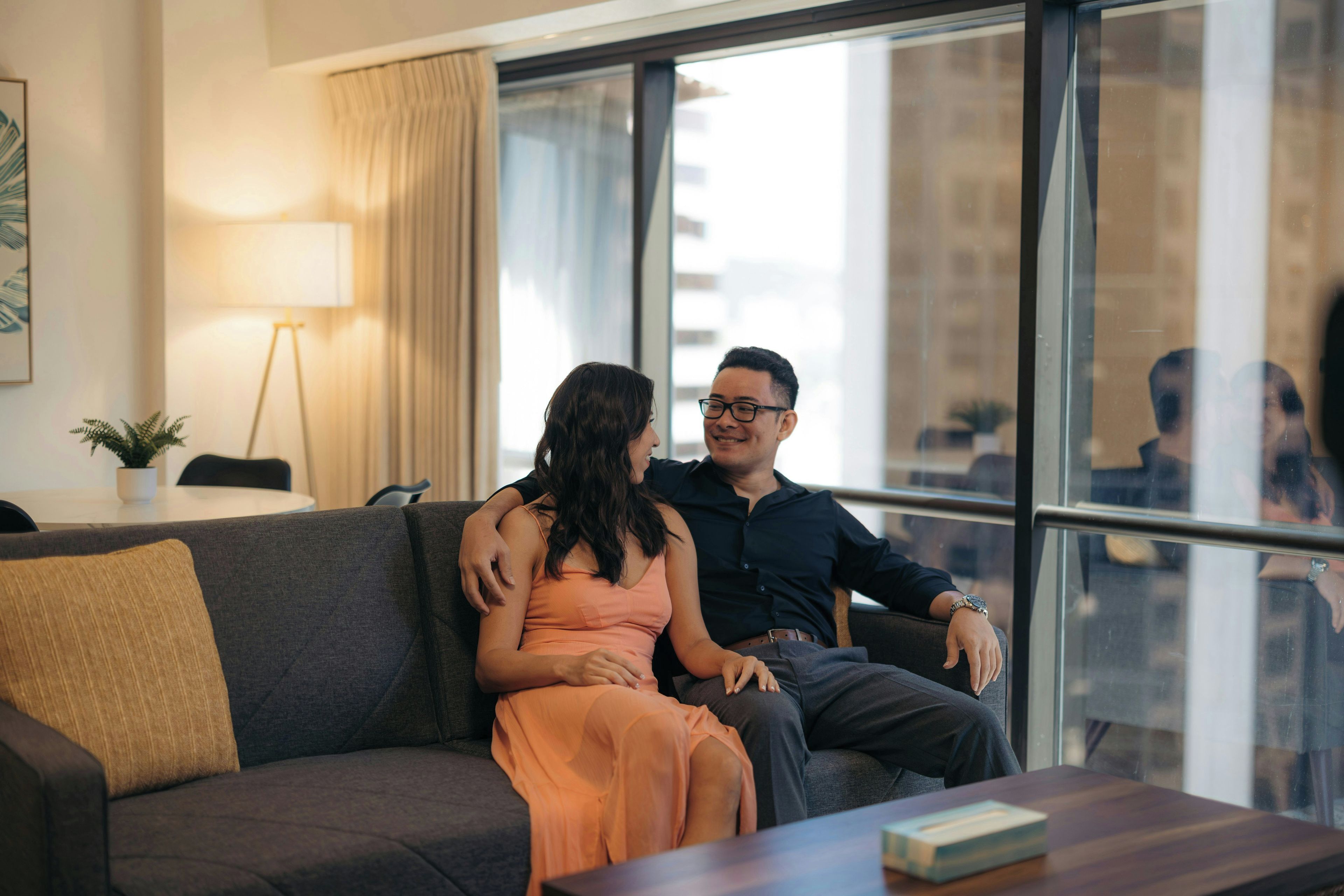 A couple sitting on a couch in a modern room with a large window, a lamp, and a view of buildings outside.