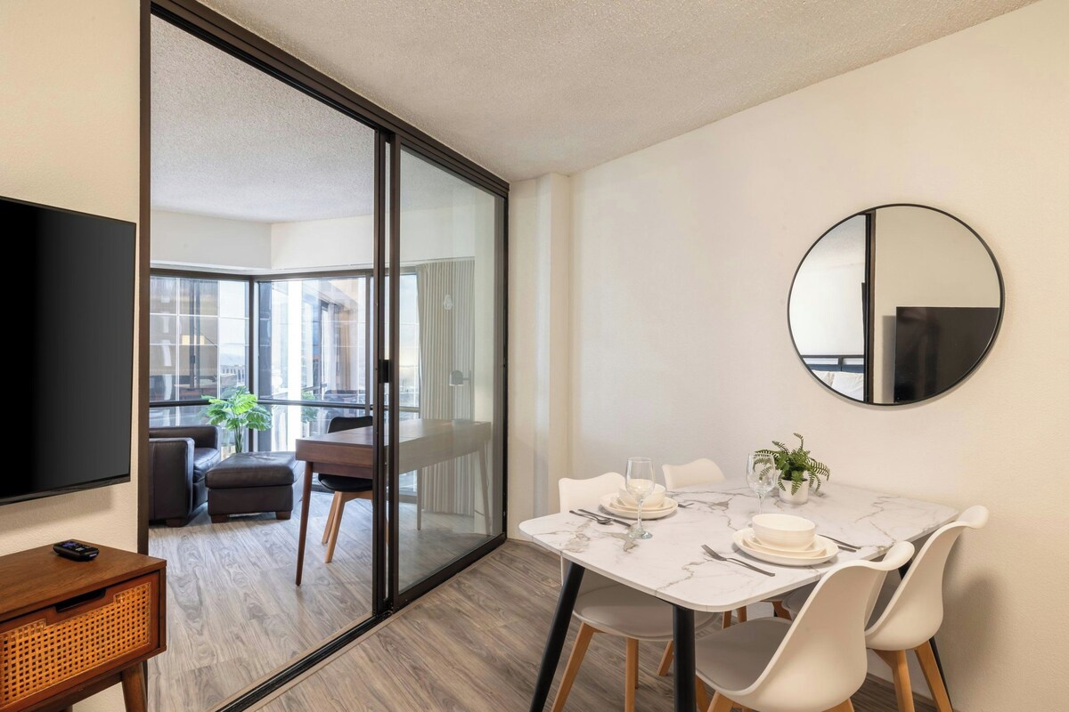 A modern dining area with a glass partition, a round mirror, and a flat-screen TV on a wooden side table.