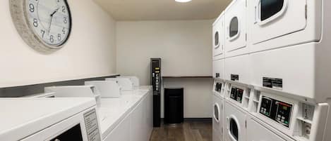 A laundry room with multiple washing machines, a clock on the wall, and a trash bin.