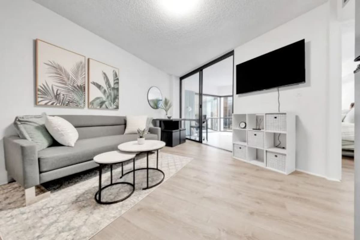 Una sala de estar moderna con un sofá gris, dos mesas redondas pequeñas, un televisor de pantalla plana montado en la pared y un estante blanco.
