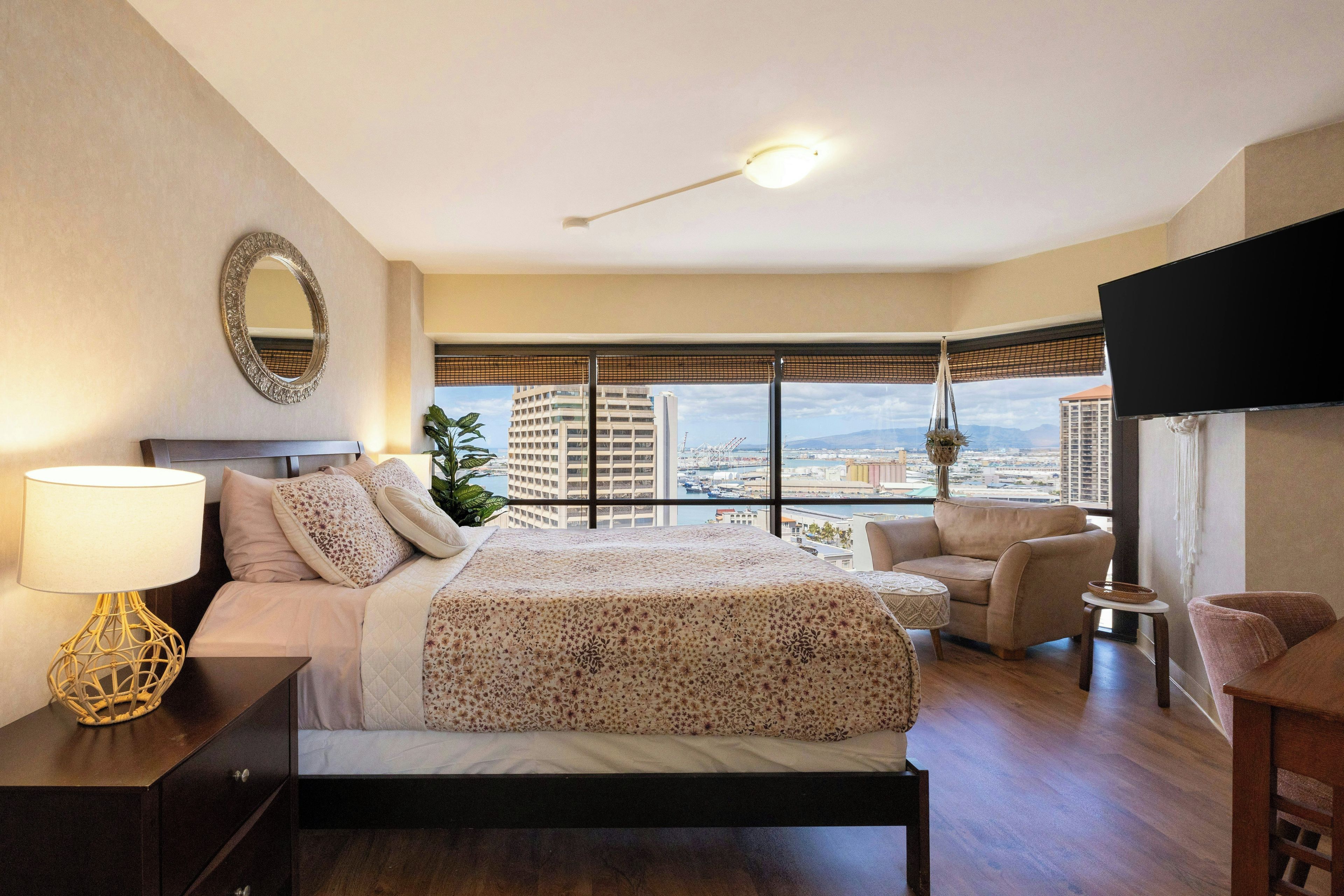 Habitación de hotel con cama, escritorio, silla, televisor y ventana grande con vista a la ciudad.