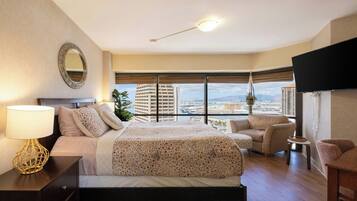 Habitación de hotel con cama, escritorio, silla, televisor y ventana grande con vista a la ciudad.